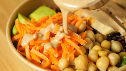 Drizzling creamy tahini dressing over vibrant vegan buddha bowl featuring shredded carrots,...