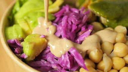 Tahini dressing cascading over colorful vegan buddha bowl, highlighting red cabbage, avocado, chickpeas, green pesto drizzle