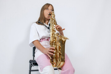 Fototapeta premium Girl playing, practicing the saxophone, playing a melody on a musical instrument, sitting on black chair on white background. Out-of-school children activities, lessons