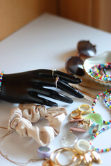 Hand figurine, sunglasses, gold jewelry, colorful bead jewelry, statement rings and various hair accessories on the white table. Selective focus.