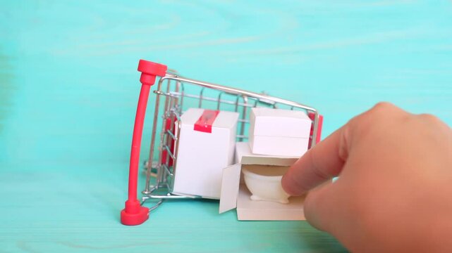 Hand positions a miniature bathtub next to an overturned shopping cart filled with small boxes, representing online ordering and delivery of bathroom supplies.
