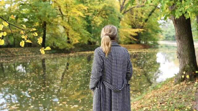 Woman in houndstooth coat stands by lake in public park. Autumn fashion clothing