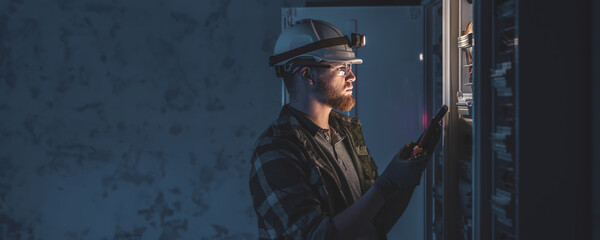 A male electrician in overalls, focused on work in switchboard with fuses, using tablet.