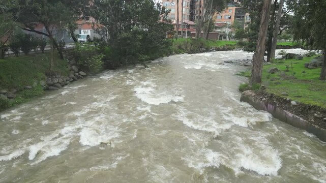 Drone footage captures the Tomebamba River winding through Cuenca, Ecuador on a cloudy day. The city skyline and surrounding hills create a dramatic backdrop.