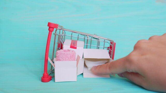 Hand positions a rubber duck on a miniature bathtub next to an overturned shopping cart filled with towels and boxes, representing playful bathroom supplies concept.