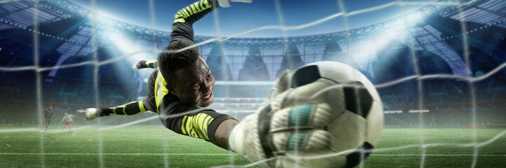 Goalkeeper in bright uniform extending his arm to catch soccer ball inches from net, with players and stadium visible in background under bright lights. Concept of sport, game, tournament, competition