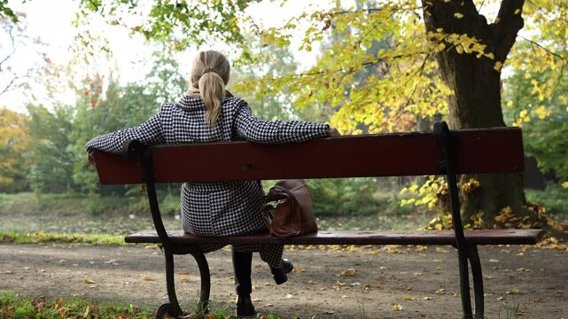 Woman relaxing on park bench in autumn and enjoying sunny day. Blond hair woman wearing houndstooth coat