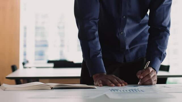 Close up man review infographics leaned at desk with papers displayed diverse charts and diagrams, hold pen write important notes on personal organizer, planning, working on data, make stats research