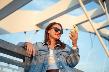 Urban woman wearing sunglasses and denim jacket holding using mobile phone in front of city bridge.