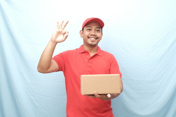 Smiling Asian delivery worker in a red uniform holding a parcel and giving an 'OK' hand gesture against a light blue background.