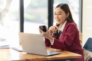 Asian woman uses smartphone while sitting at table with laptop computer at office She writes notes on document graphs. female financial accountant with financial growth statistics chart