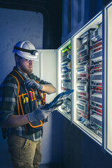 Electrician in overalls, focused on work in switchboard with fuses, using tablet.