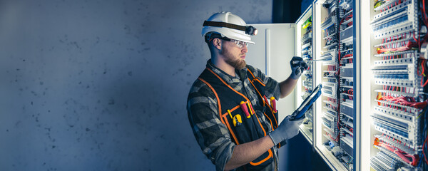 A male electrician in overalls, focused on work in switchboard with fuses, using tablet.