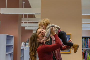 A joyful family scene with a mother lifting her smiling toddler in the air as the father lovingly looks on in a cozy library setting. Cleveland, Ohio, USA