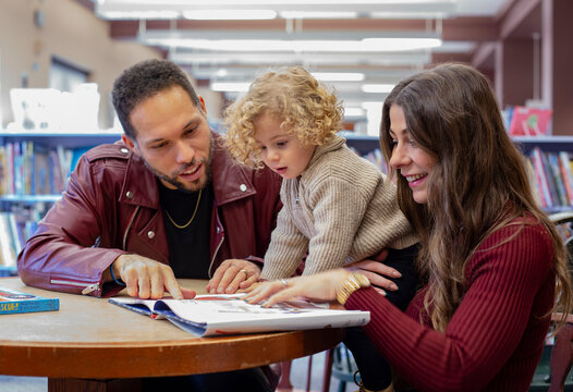 A family enjoys reading together in a library with shelves of books in the background. Cleveland, Ohio, USA