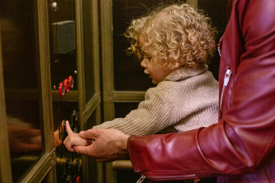 Curly-haired child learning to press an elevator button with the help of an adult's guiding hand. Cleveland Public Library, Ohio, USA