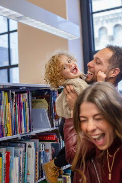 Joyful family having fun in a library with a laughing child being held by her father while the mother laughs along. Cleveland, Ohio, USA