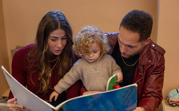 Parents and their young child are engaged in reading a large picture book together, with focus and enjoyment evident in their interaction. Cleveland, Ohio, USA