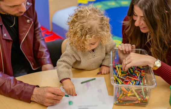 A toddler with curly hair is coloring with crayons at a table, flanked by a man and a woman who are observing and assisting. Cleveland, Ohio, USA