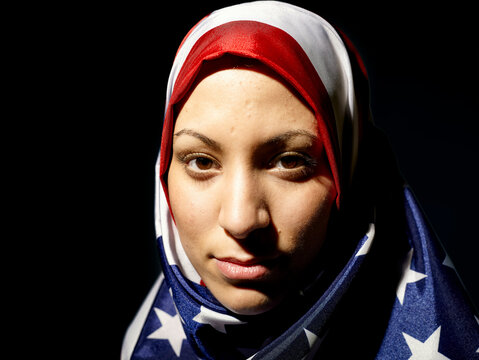A confident woman wearing a hijab with the American flag pattern against a dark background.