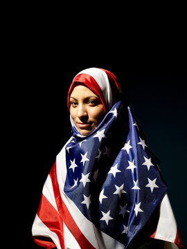 Woman draped in an American flag hijab looking at the camera with a subtle smile against a dark background.