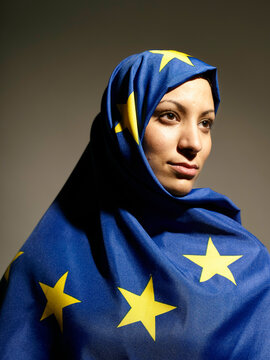 A woman draped in a European Union flag themed hijab looks thoughtfully to the side against a gray background