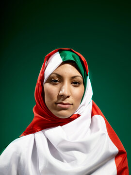 A woman draped in a flag with a red, white, and green design stands against a green background, her expression serene and respectful.