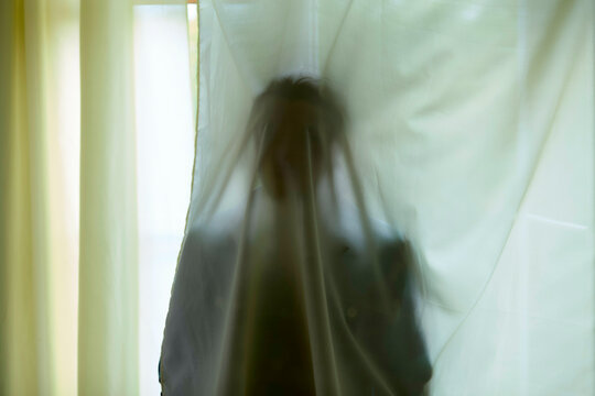 A blurred silhouette of a person behind white curtains exhibiting an abstract and ethereal appearance.