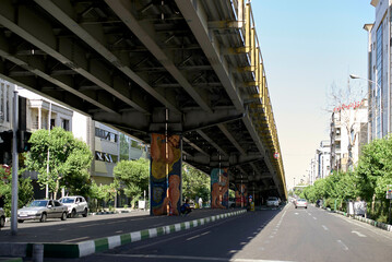 Urban scene with cars driving under an overpass adorned with colorful art installations on the pillars in a sunny day, Teheran, Iran