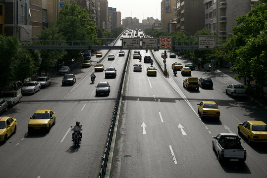 A bustling city street with multiple lanes of traffic including cars, buses, motorcycles, and a pedestrian overpass in the background, Teheran, Iran