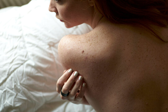 Close-up of a woman with red hair laying on a bed, showing her bare shoulder and arm with a subtle touch