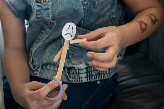 A girl holds a handmade stick figure with a sad face drawn on it while sitting down, showcasing a DIY craft project, Netherlands