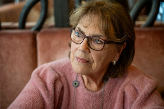 Older woman with glasses and a thoughtful expression sits at a table in a cozy setting, wearing a pink sweater, Netherlands - Powered by Adobe