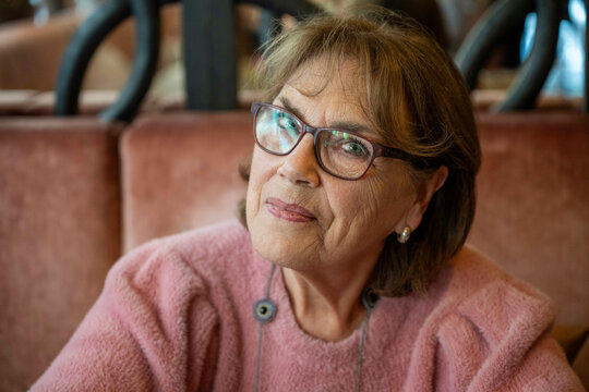 Elderly woman wearing glasses and a pink sweater sits comfortably indoors, exuding warmth and wisdom, Netherlands