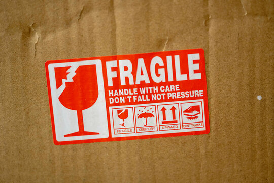 Red and white fragile label on a cardboard box, featuring icons and text for handling instructions: fragile, keep dry, upward, don't trample