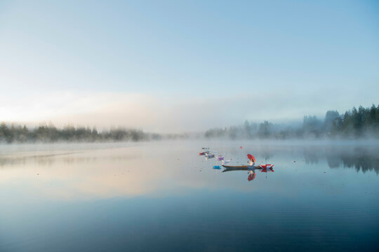 A serene, misty lake reflects the sky in the early morning light, with colorful kayaks floating in the calm water, surrounded by a line of dense trees at the horizon.