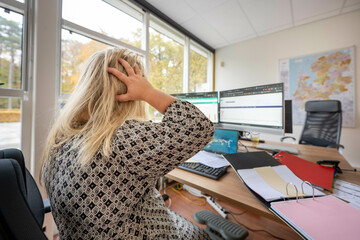Person with long blonde hair sits at an office desk, facing away, with hands on head. Multiple computer screens display graphs and data, Molenhoek, Netherlands
