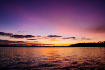 A breathtaking sunset over a calm lake with a silhouette of a tree-lined shore. The vibrant sky transitions from deep purple to soft orange and pink hues, Norwegian Point, WA, USA