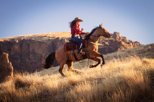 A woman in a red shirt and cowboy hat rides a galloping horse through sunlit, grassy plains against a backdrop of rugged cliffs, Idaho, USA