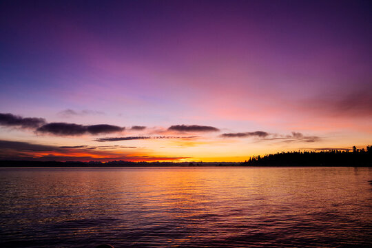 A breathtaking sunset over a calm lake with a silhouette of a tree-lined shore. The vibrant sky transitions from deep purple to soft orange and pink hues, Norwegian Point, WA, USA