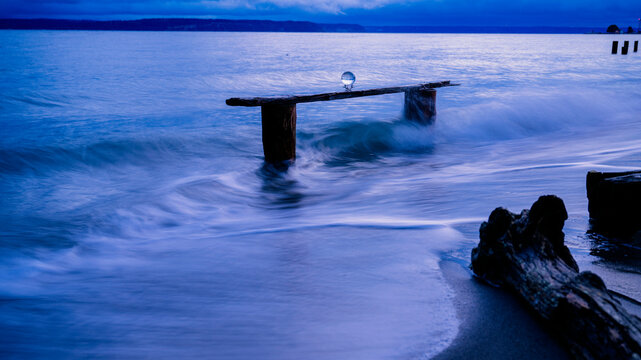 A serene beach scene with gentle waves splashing against a wooden structure, topped with a glass sphere, under a deep blue twilight sky, Norwegian Point, WA, USA
