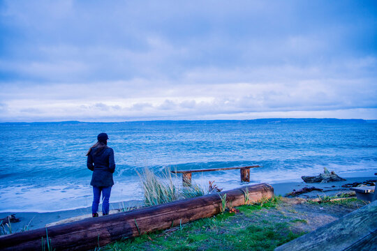 A woman in a dark coat and hat stands on a beach, looking out at the expansive ocean under a cloudy sky. Driftwood, Norwegian Point, WA, USA