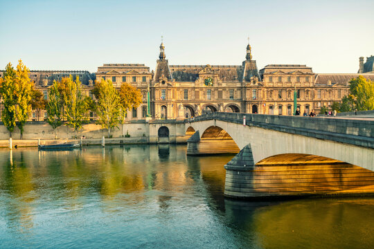 Elegant historical building of the Louvre Museum and bridge over a calm Seine river at sunset, with clear skies and warm sunlight bathing the scene, Paris, France