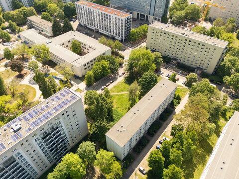 Aerial view of a modern building with a solar-paneled roof beside traditional rooftops surrounded by lush green trees and parked cars, Berlin, Germany