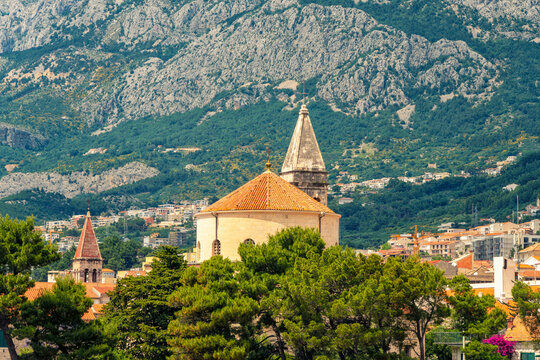 Ancient  Franciscan Monastery of the blessed virgin Mary with a steeple stands before a mountain with lush greenery under a cloudy sky, Makarska, Croatia