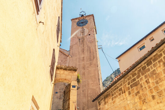 Sunlit old clock tower  of the Church of the Holy Spirit in Omis, between historic stone buildings under a clear blue sky, Croatia
