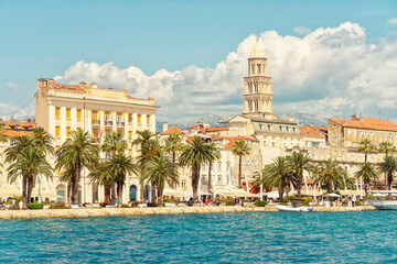 Palm trees line the waterfront promenade in front of historic buildings and St Domnius Cathedral bell tower, under a clear blue sky with fluffy clouds, Split, Croatia