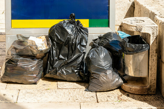 Black garbage bags piled beside a stone waste container on a sunny day with an informational sign in the background, Split, Croatia