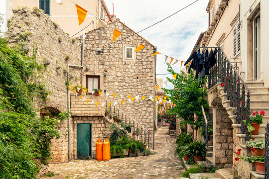 A quaint cobblestone alley adorned with flags and plants between traditional European stone houses, with clothes hung out to dry on a bright day, Mali Ston, Croatia