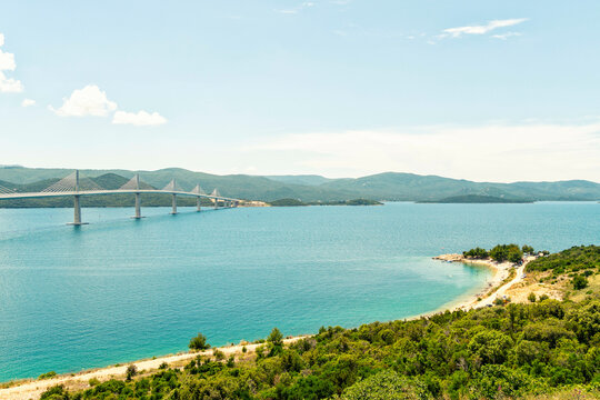 Coastal landscape featuring a long suspension Peljesac Bridge spanning across a blue bay with a lush green shoreline under a clear sky, Peljesac, Croatia